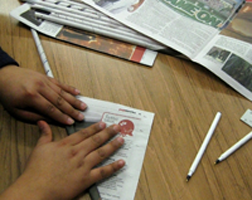 A person is writing on paper with pens and newspapers.